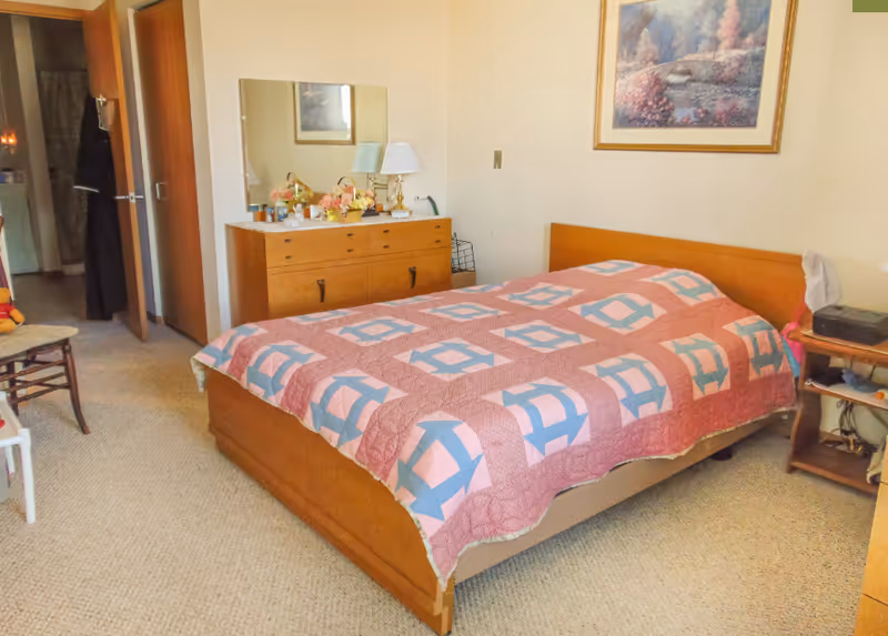 A bedroom with a wooden bed frame and a quilted bedspread in pink, blue, and white geometric patterns. There is a wooden dresser with a mirror above it, decorated with a lamp, flowers, and various small items. A framed painting hangs on the wall above the bed. The room has beige carpet and light-colored walls. A chair and a small side table with a printer are also visible. An open door leads to another room or hallway.