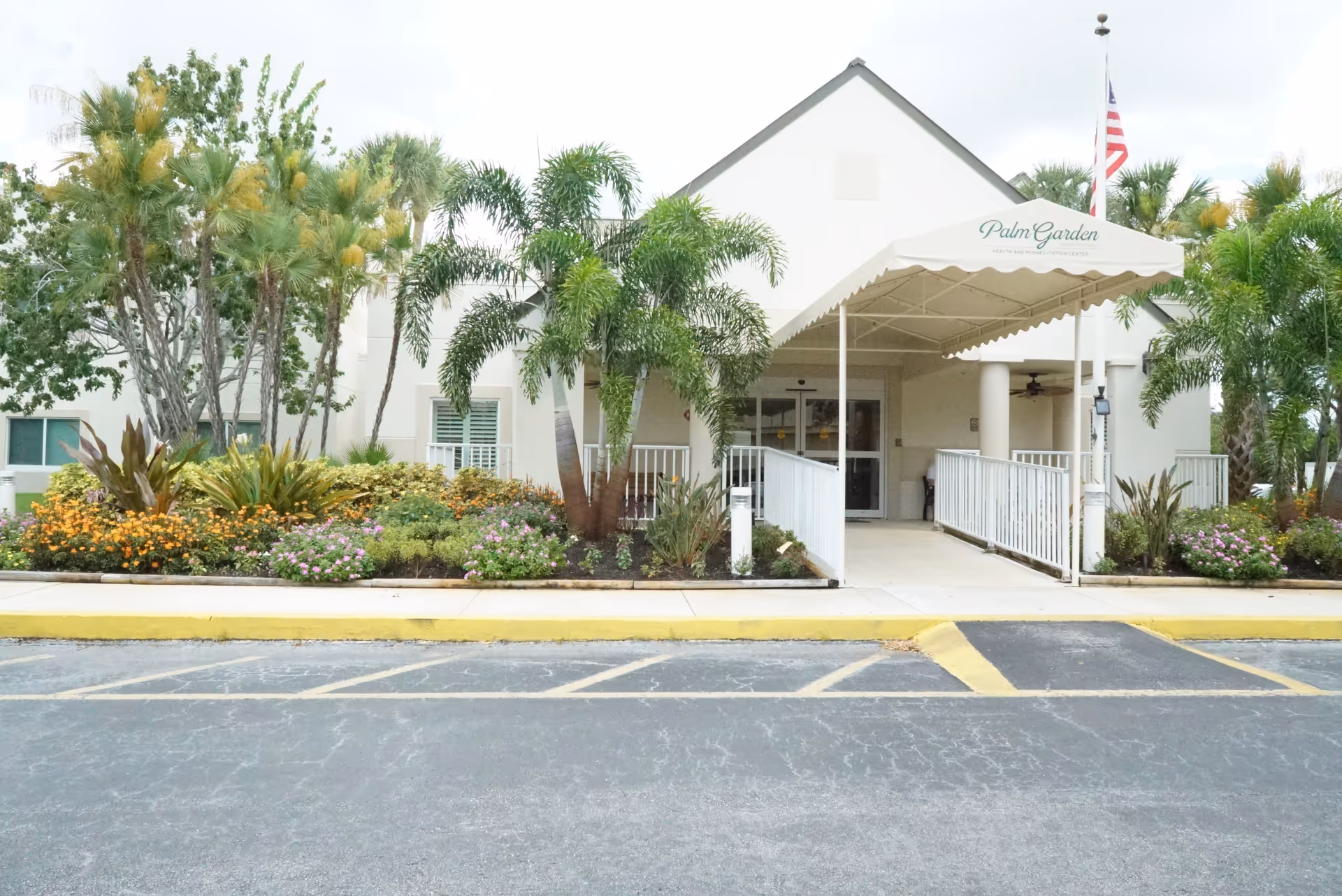 Entrance of Palm Garden of Port St Lucie with a covered canopy, ramp, palm trees and landscaped flowerbeds.