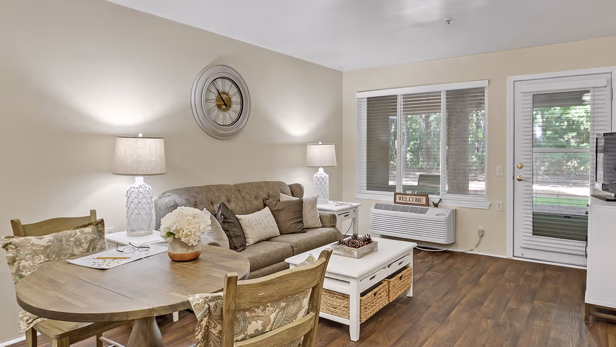 Cozy living room with a sofa, dining table, and large windows showing greenery outside.