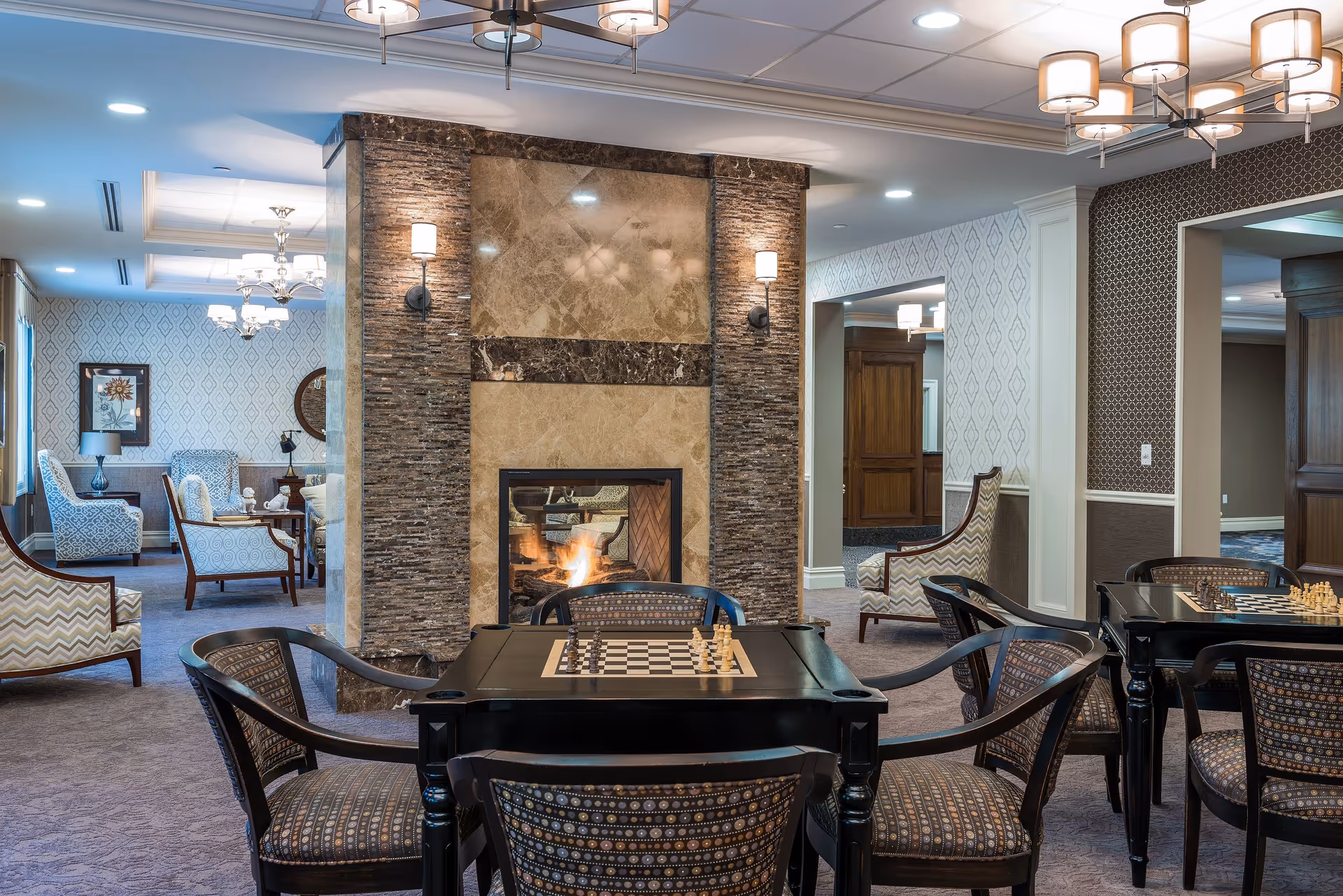 A cozy senior living facility common area with a central fireplace surrounded by stone and marble. In front of the fireplace are two black tables with built-in chessboards and patterned chairs. The room features soft lighting from ceiling fixtures and wall sconces. In the background, there are additional seating areas with upholstered armchairs and decorative wallpaper.