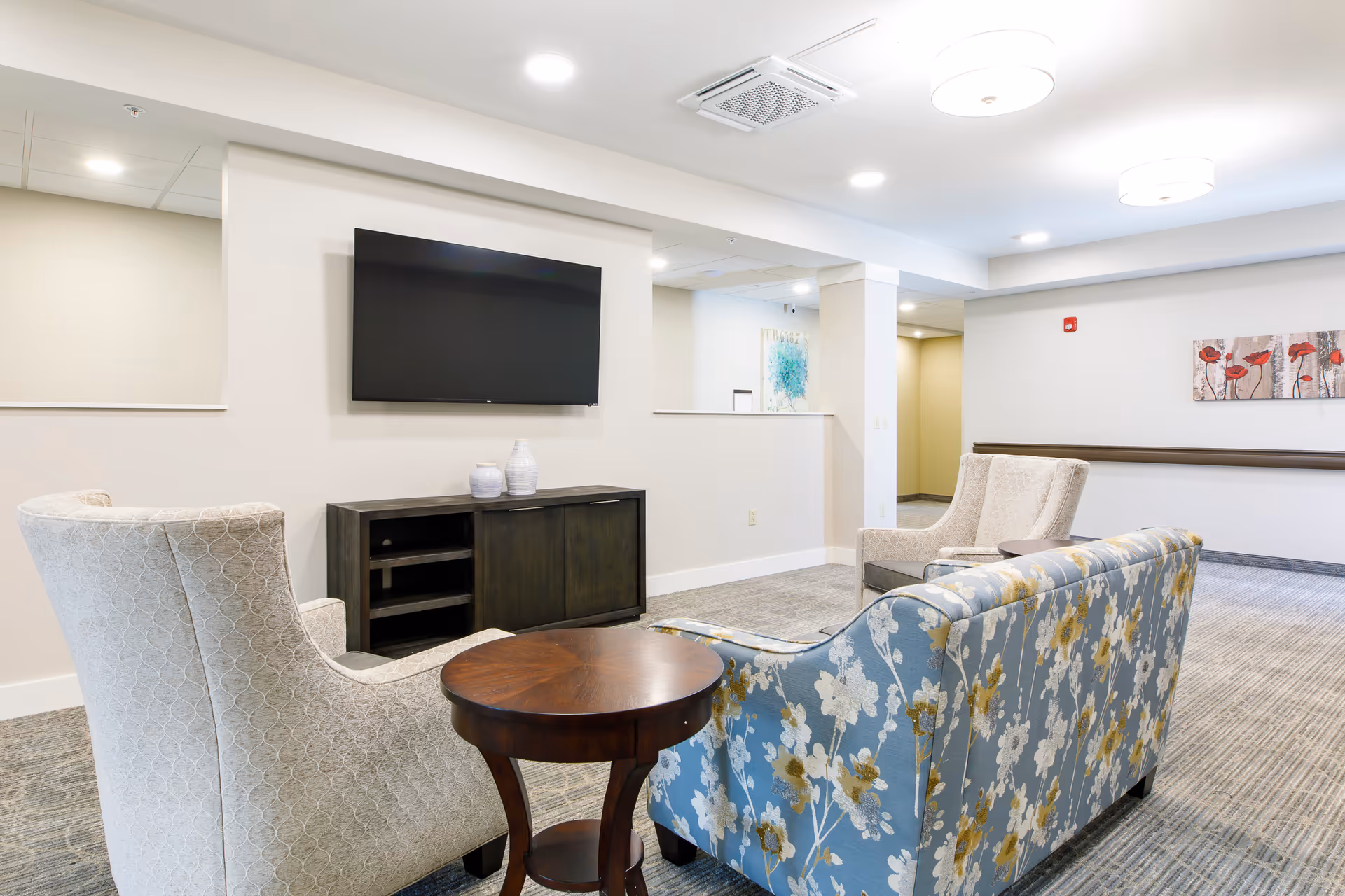 Bright communal lounge with a floral sofa, two armchairs, a wall-mounted TV and a small side table.