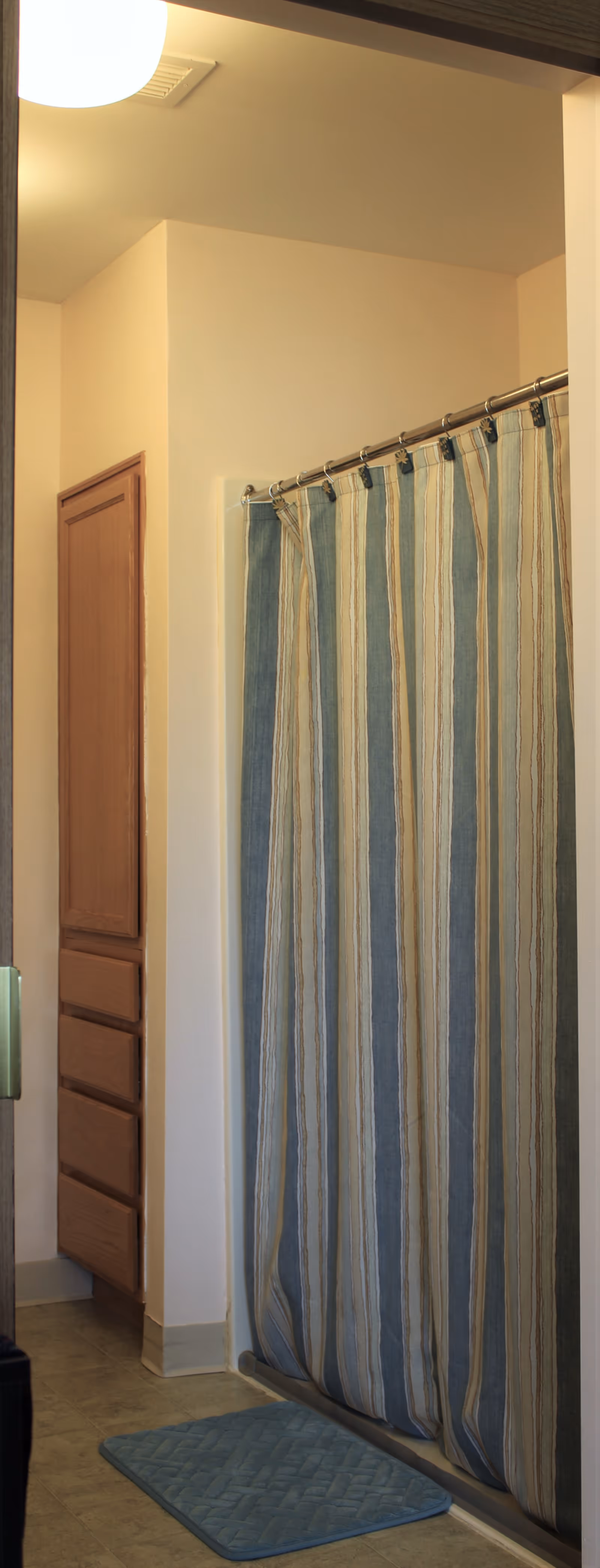 A bathroom with a striped shower curtain in shades of blue, beige, and white hanging from a metal rod. There is a blue bath mat on the floor in front of the shower. To the left, there is a wooden cabinet with drawers and a door. The walls are painted light beige, and a round ceiling light is visible.