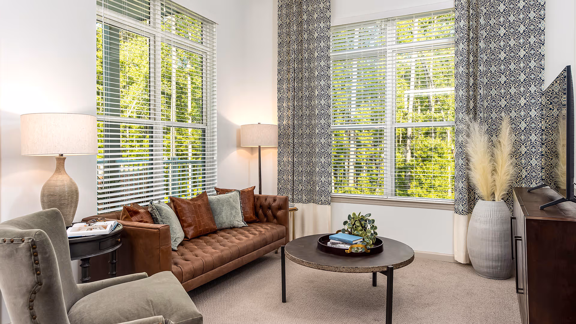 A bright living room with large windows covered by white blinds and patterned curtains. The room features a brown tufted leather sofa with multiple cushions, a round coffee table with books and a plant, a gray upholstered armchair, two lamps with beige shades, and a large vase with pampas grass next to a flat-screen TV on a wooden stand.
