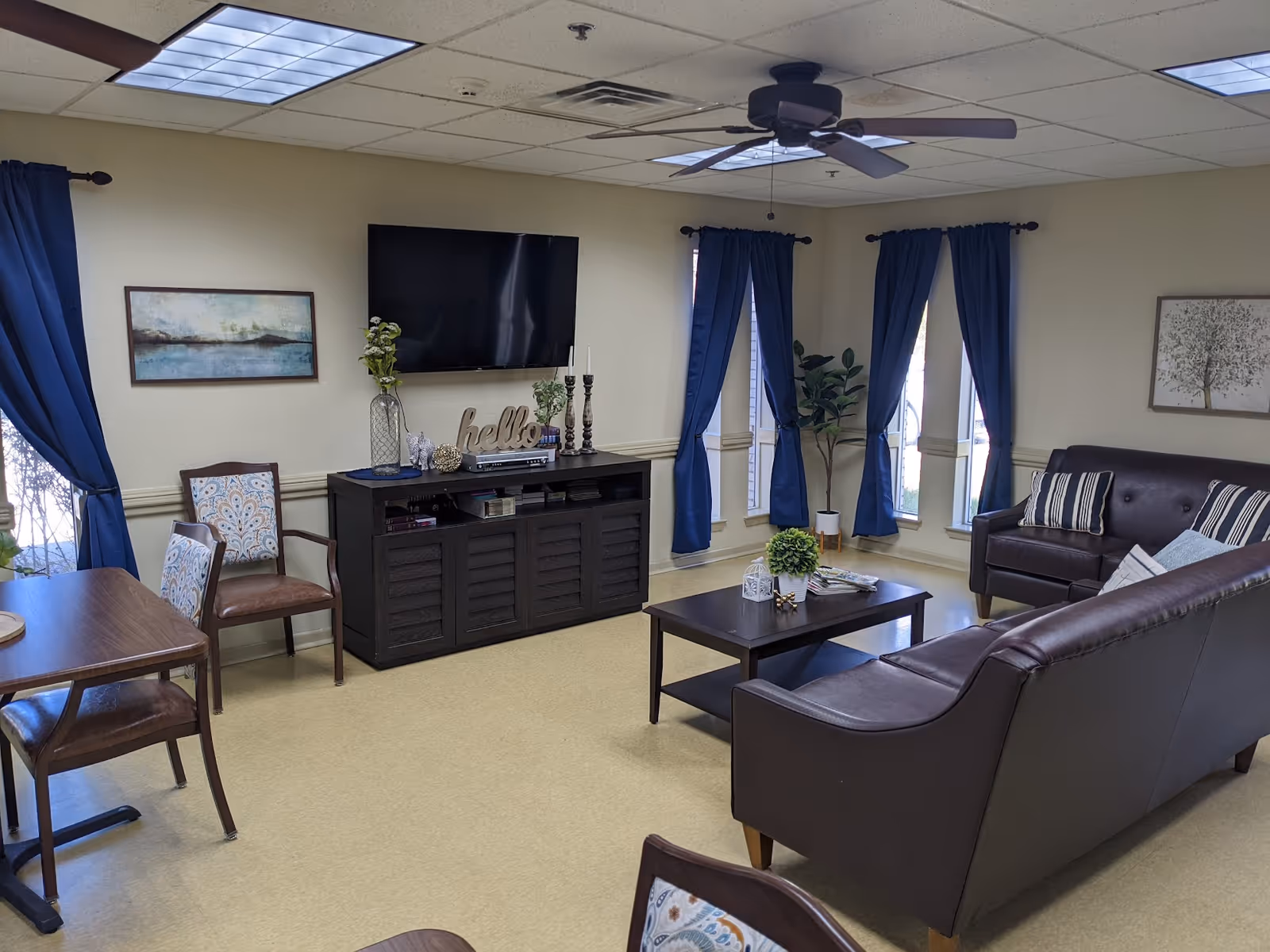 A cozy living room area with two dark brown leather sofas, a wooden coffee table with decorative plants and magazines, a TV mounted on the wall above a black media console, blue curtains on windows, a ceiling fan, and a small dining table with chairs.