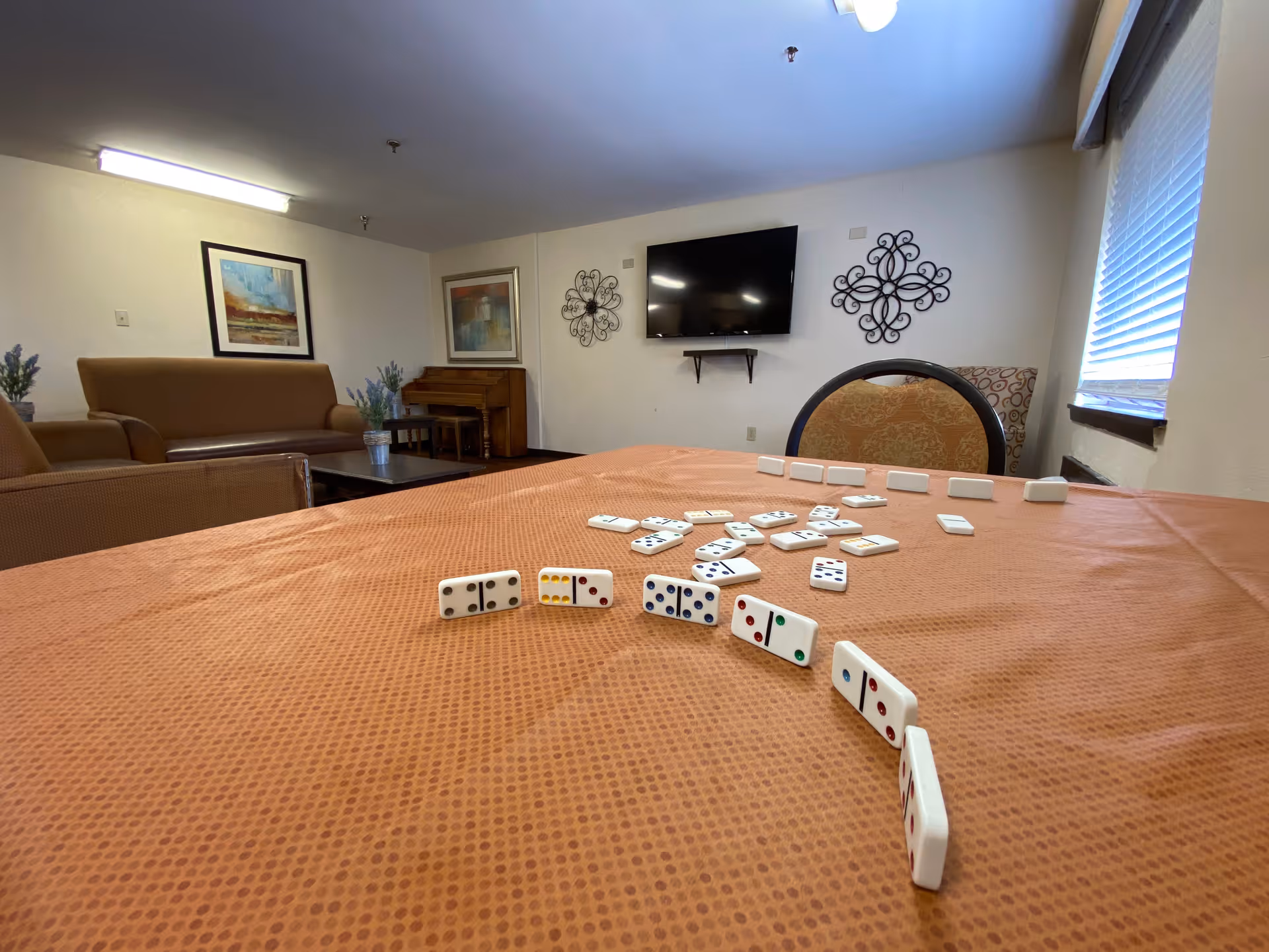 A cozy common room with a table covered in an orange tablecloth scattered with dominoes. In the background, there are brown sofas, a piano, framed artwork on the walls, a flat-screen TV mounted on the wall, decorative wall hangings, and a window with blinds letting in natural light.