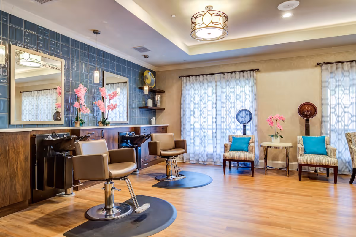 A bright and modern salon area with two brown salon chairs in front of sinks and mirrors mounted on a blue tiled wall. There are pink flowers on the counter, two armchairs with blue cushions, a small round table with pink flowers, and two vintage hair dryers near large windows with patterned curtains. The floor is wooden, and the ceiling has a decorative light fixture.