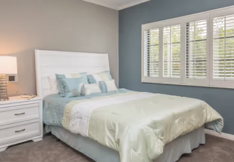 A neatly made bed with light green and blue bedding and multiple pillows in a bedroom with gray and blue walls. There is a white nightstand with a lamp and a phone on the left side of the bed, and a window with white plantation shutters on the right wall.