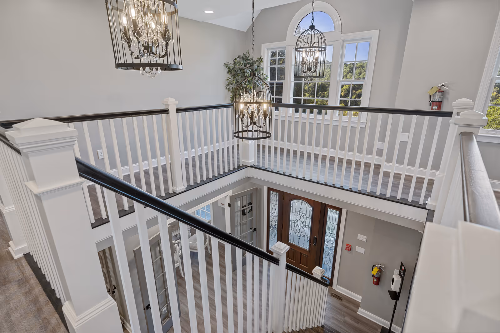 Interior view of a senior living facility showing a two-story entryway with white railings and dark handrails. There are two hanging chandeliers with cage-like designs, a large arched window letting in natural light, and a wooden front door with decorative glass panels. The walls are painted light gray and the flooring is wood-style.