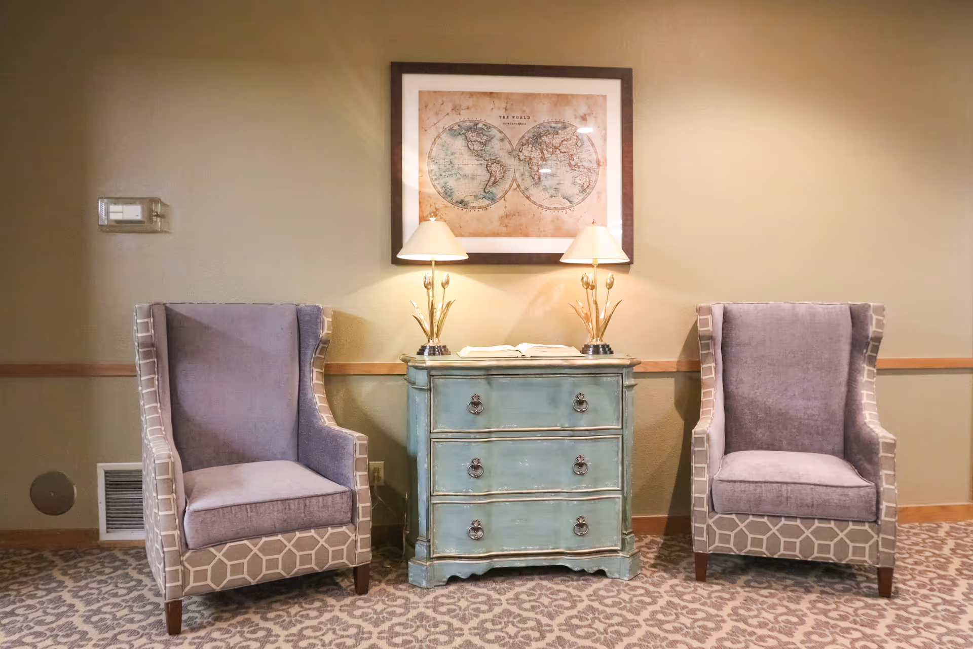 Two upholstered armchairs with patterned sides are placed on either side of a distressed blue chest of drawers with three drawers. On top of the chest are two table lamps with beige shades and a book. Above the chest hangs a framed vintage world map. The room has beige walls and patterned carpet flooring.