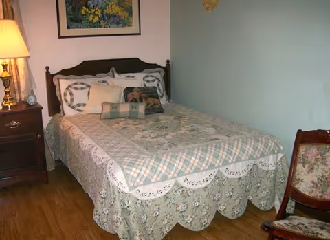 A cozy bedroom with a wooden bed frame, a bed covered with a floral and plaid quilt, several decorative pillows, a wooden nightstand with a lamp and an alarm clock, a floral upholstered chair, and a framed floral painting on the wall.