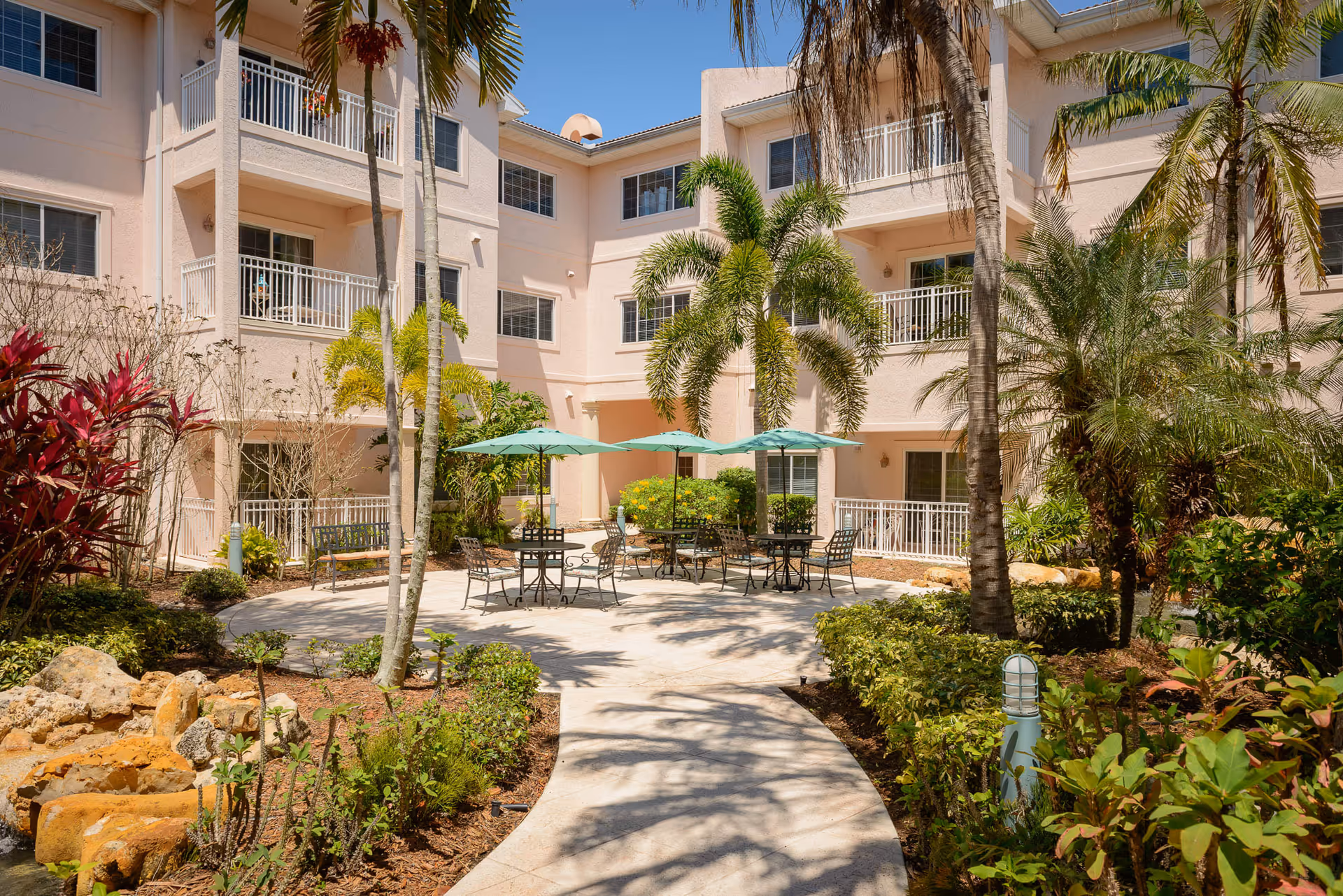 Sunlit courtyard with patio tables and umbrellas surrounded by palm trees and a three-story residential building with balconies.