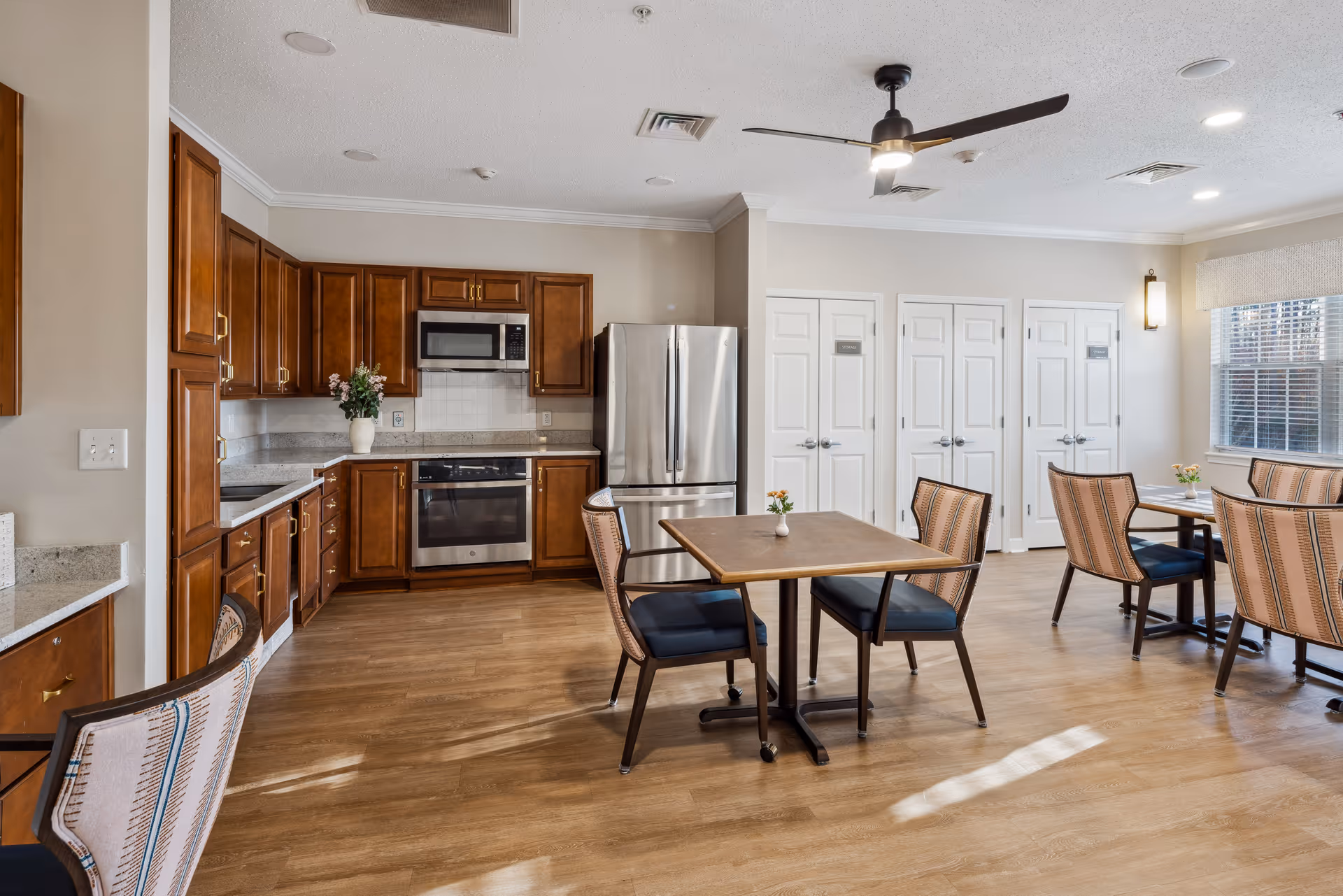 Communal dining area with wooden tables and chairs adjacent to a kitchen with stainless steel appliances and wooden cabinets.