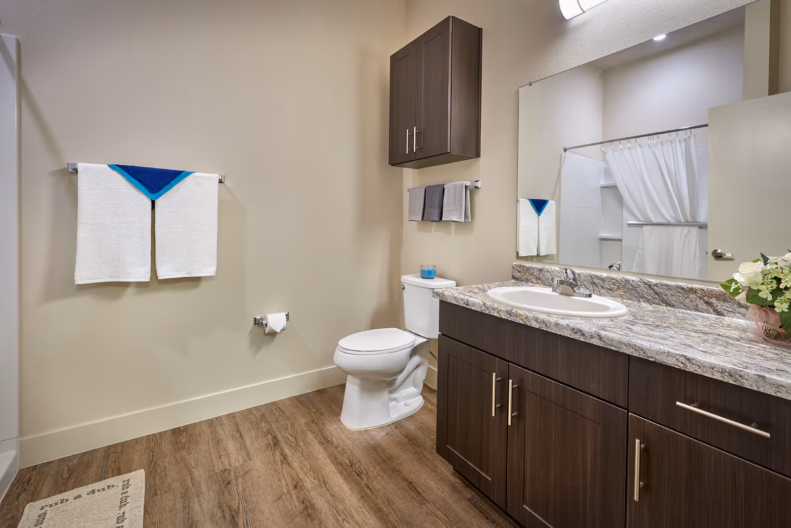 A clean and modern bathroom with wood-look flooring, a white toilet, a granite countertop with a sink, dark wood cabinets, a large mirror, a wall-mounted cabinet, and neatly hung towels. A white shower curtain is visible in the reflection of the mirror.