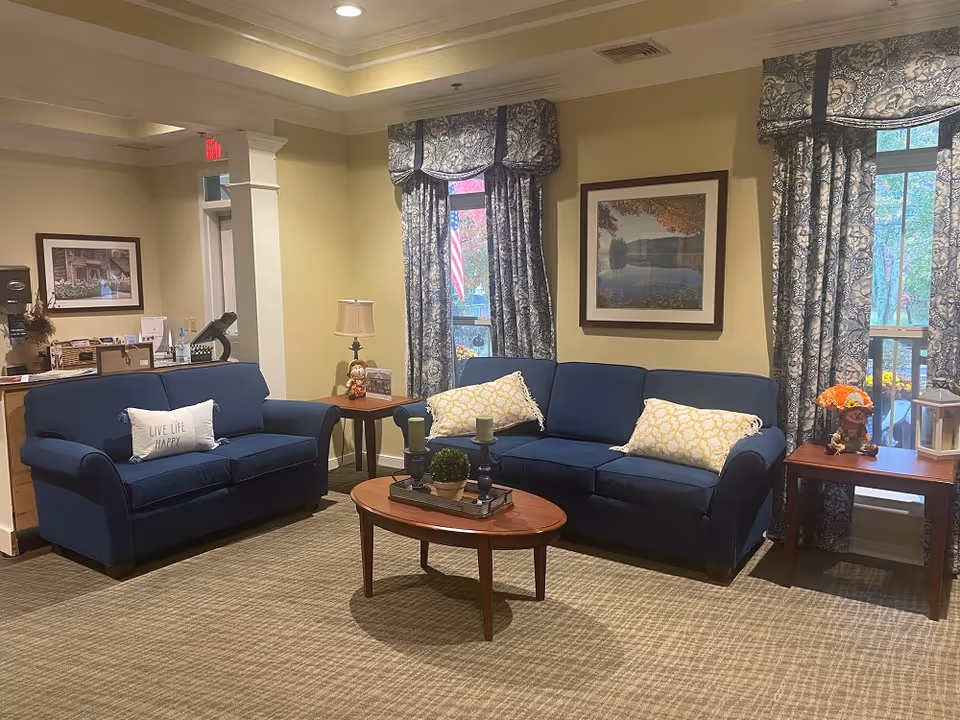Cozy seating area with two blue sofas, a wooden coffee table, side tables, framed artwork, and patterned curtains.