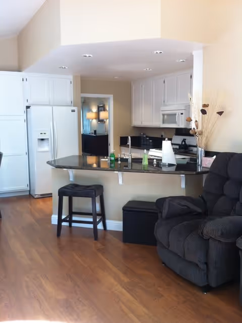 Open-plan kitchen with a granite breakfast bar, white cabinets, a refrigerator, and an adjacent recliner.