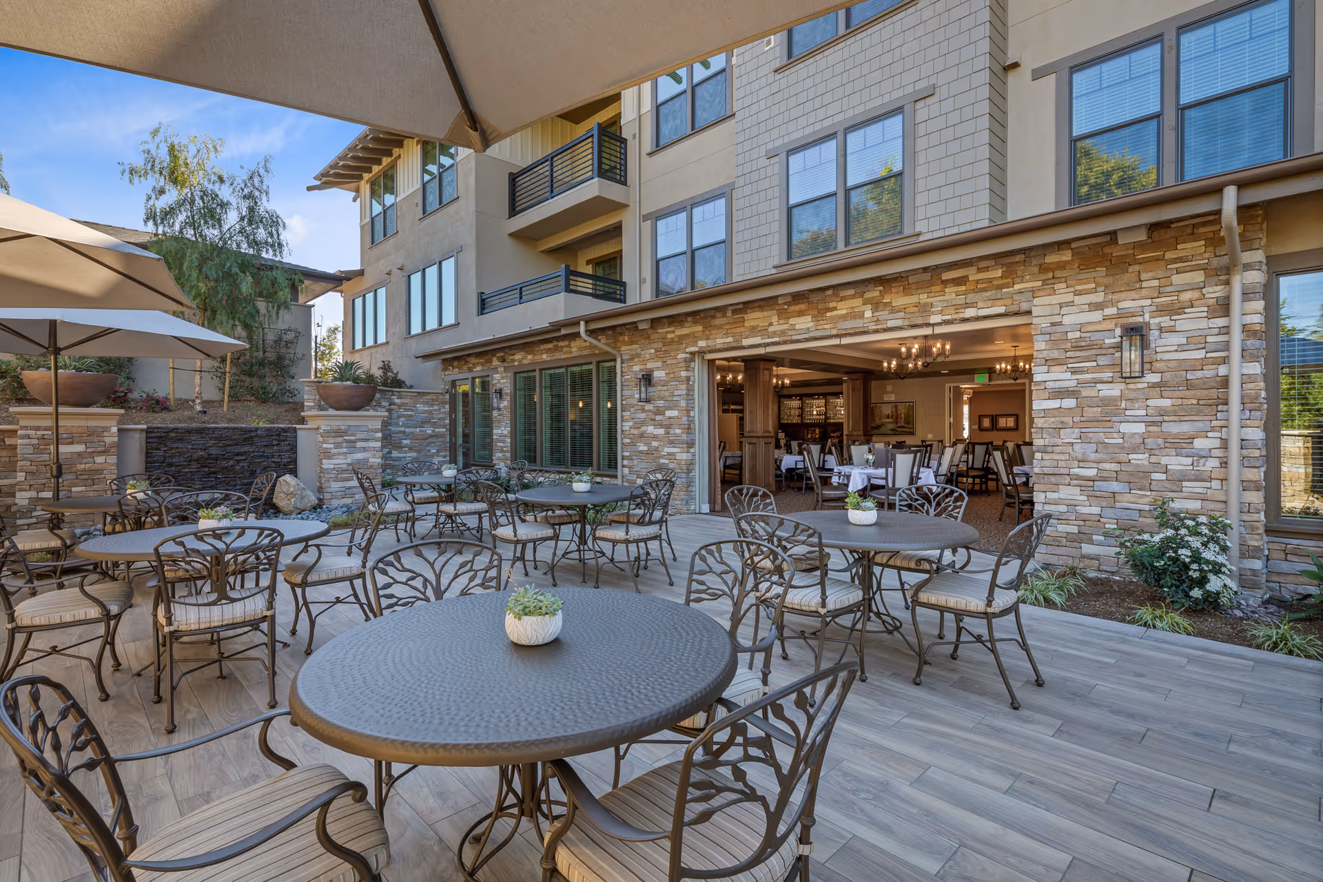 Outdoor patio with round metal tables and chairs in front of a stone-clad building opening to an indoor dining area.