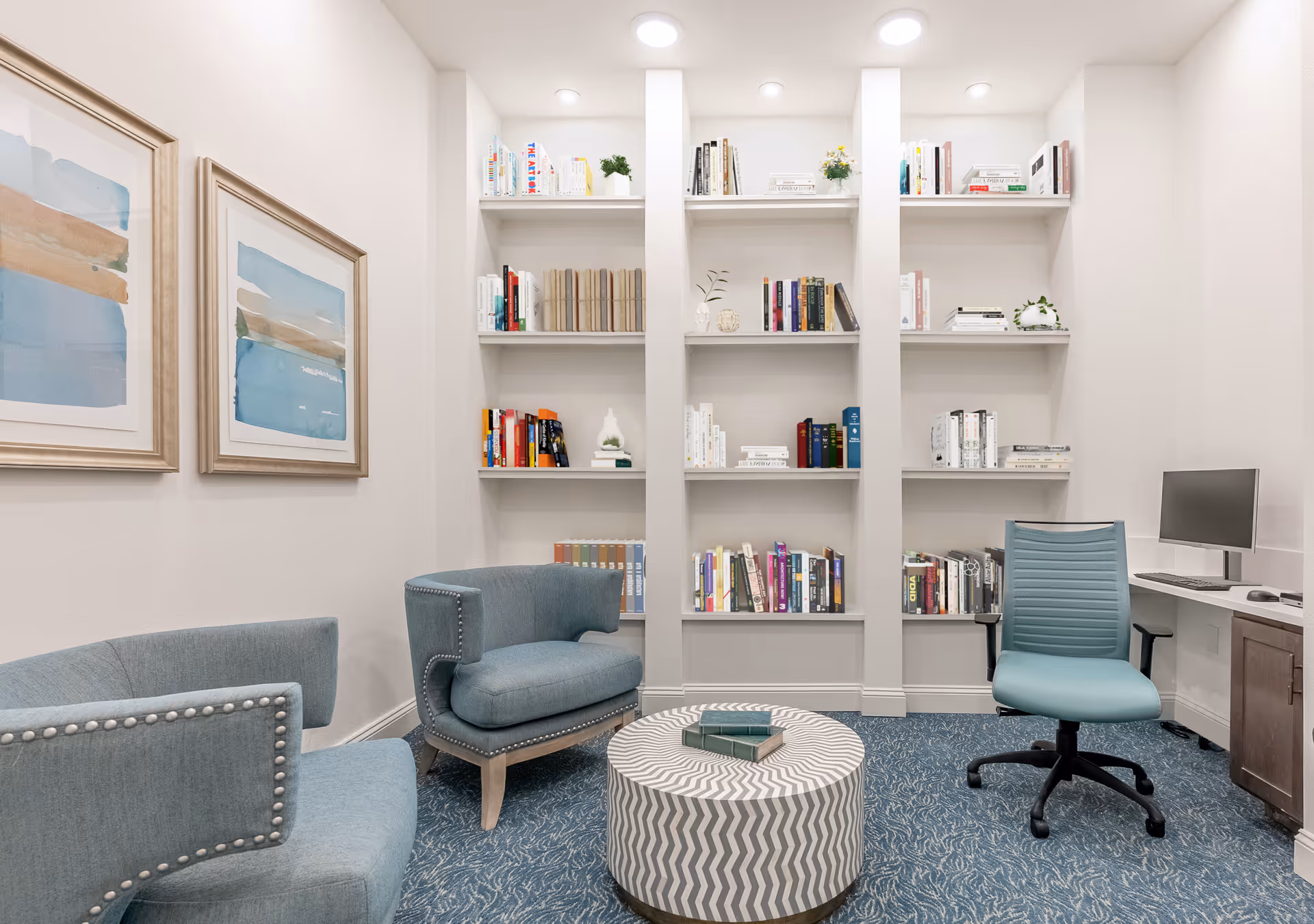 A cozy reading and work area with built-in white bookshelves filled with books and decorative items. The room features two blue upholstered chairs with nailhead trim, a round patterned ottoman with books on top, and a blue office chair in front of a desk with a computer monitor. The walls are light-colored with two framed abstract paintings, and the floor is covered with a blue patterned carpet.