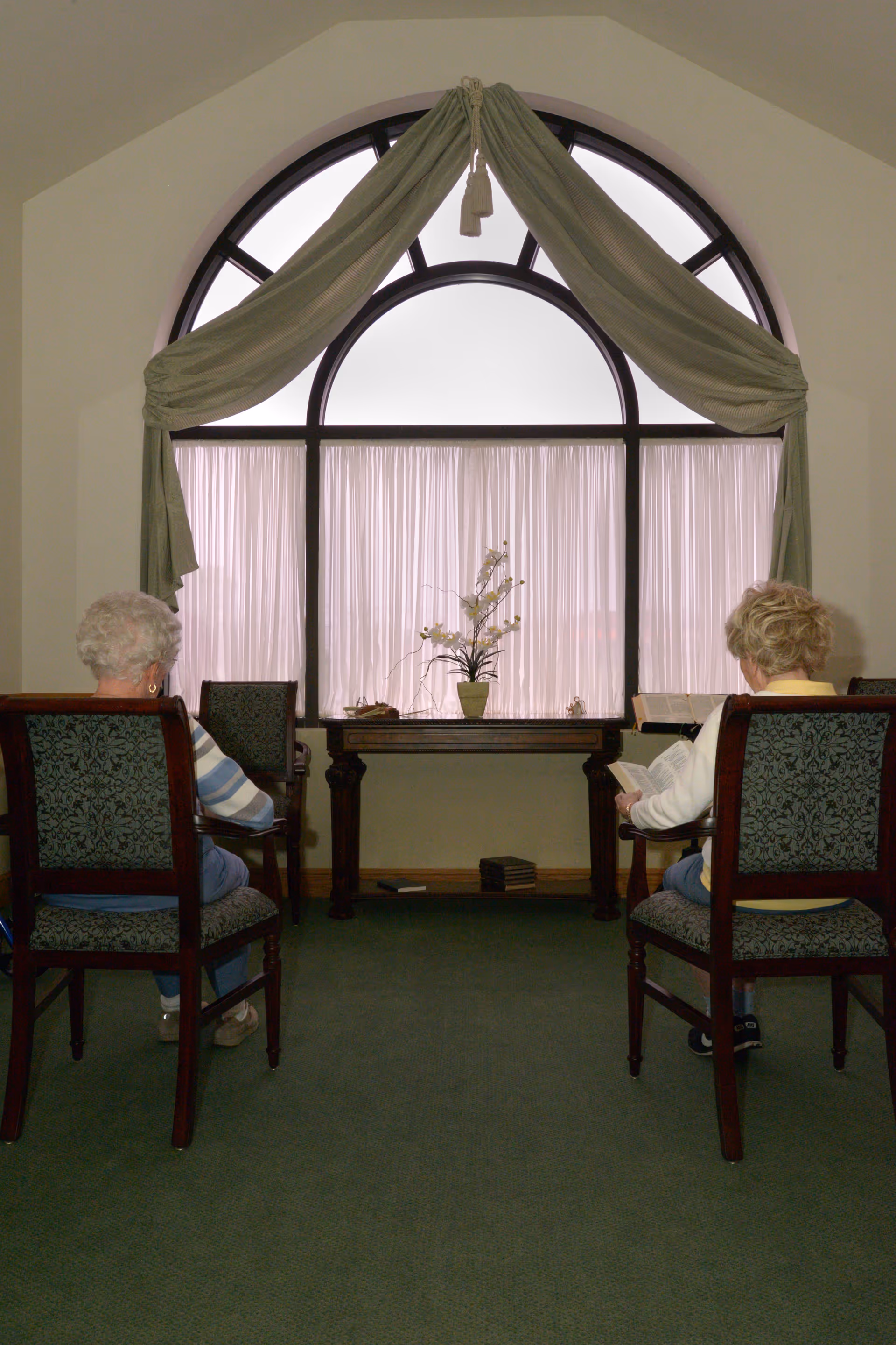 Two elderly women sit in chairs facing a large arched window with a small table and potted plant between them.