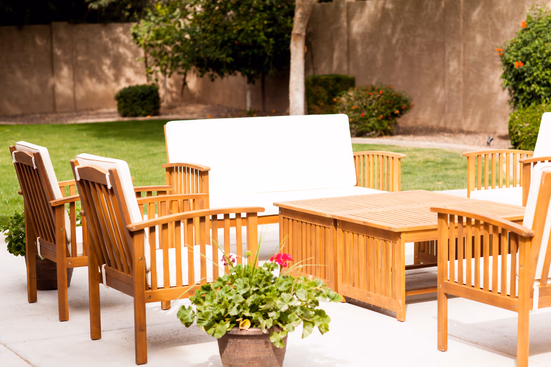 Outdoor patio area with wooden chairs and a loveseat featuring white cushions arranged around a wooden coffee table. There are potted plants with green leaves and flowers in the foreground, and a grassy lawn with bushes and a tree in the background.
