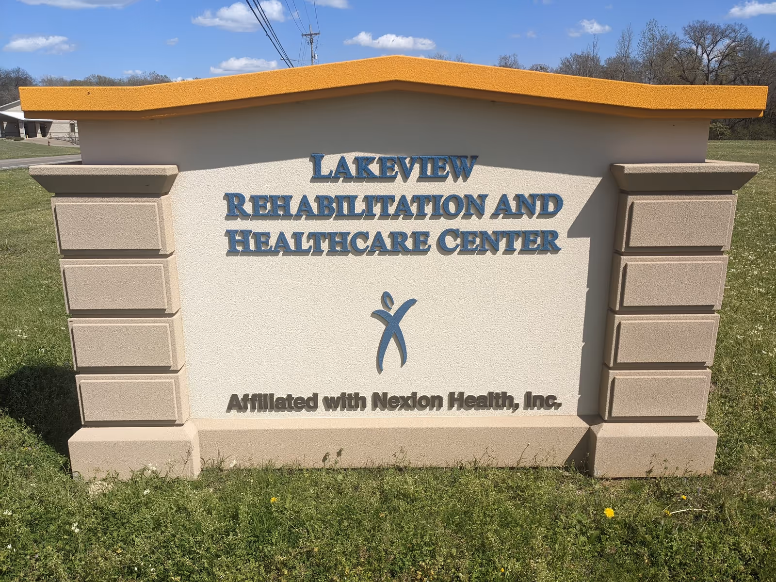 Outdoor sign for Lakeview Rehabilitation and Healthcare Center with a blue logo and text indicating affiliation with Nexlon Health, Inc., set on a grassy area under a blue sky with some clouds.