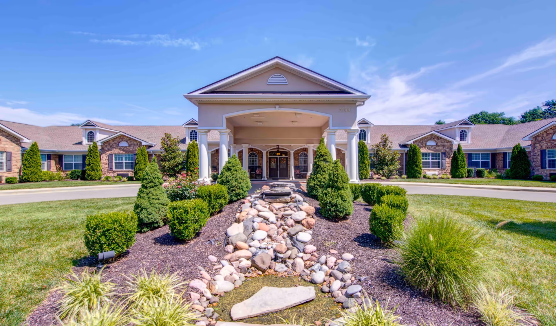 Front exterior view of Addington Place of Prairie Village, featuring a covered entrance with white columns, a stone pathway with decorative rocks, neatly trimmed bushes, green lawn, and a clear blue sky.