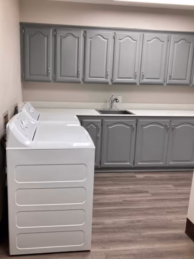 Laundry room with two top-loading machines, gray cabinets, a countertop and a sink.