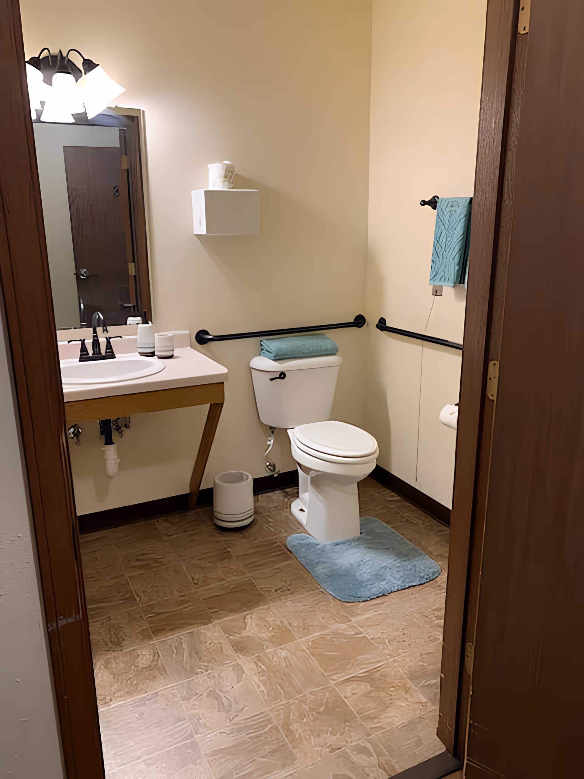 Accessible bathroom with a toilet, sink and mirror, grab bars, and teal towels and bath mat.