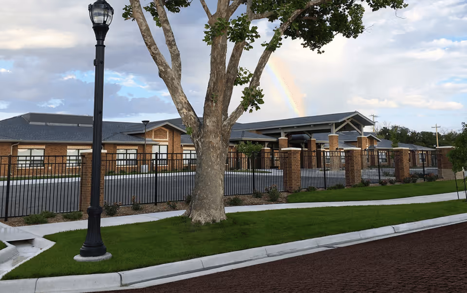 Exterior view of a senior living facility with a brick building, black metal fence, a large tree, a street lamp, and a rainbow in the sky.