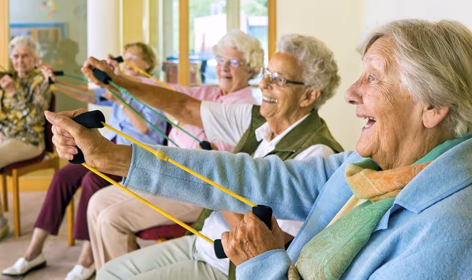 A group of elderly women sitting in chairs indoors, smiling and exercising with yellow resistance bands stretched in front of them.