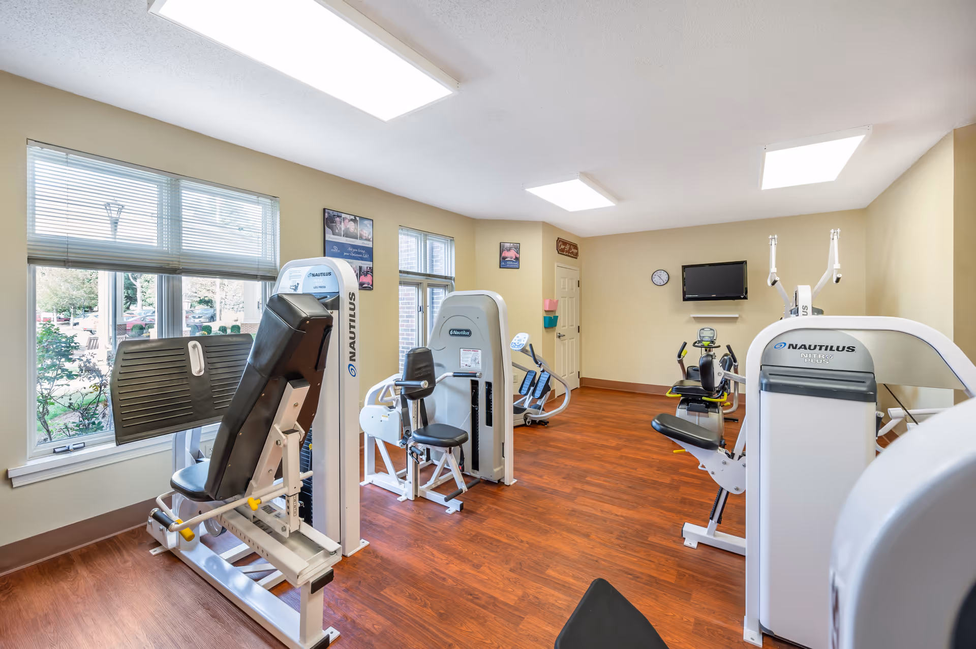 A bright fitness room with several Nautilus exercise machines arranged on a wooden floor. Large windows on the left wall let in natural light, and a wall-mounted TV and clock are visible on the far wall. The walls are painted beige, and there are a few framed pictures and a door in the background.