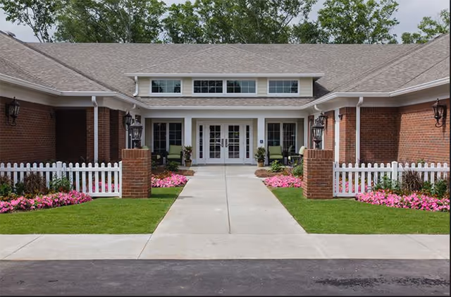 Front exterior view of Kemp Meadows Assisted Living facility showing a brick building with a white picket fence, green lawn, pink flowers, and a concrete walkway leading to double glass doors under a covered entrance.