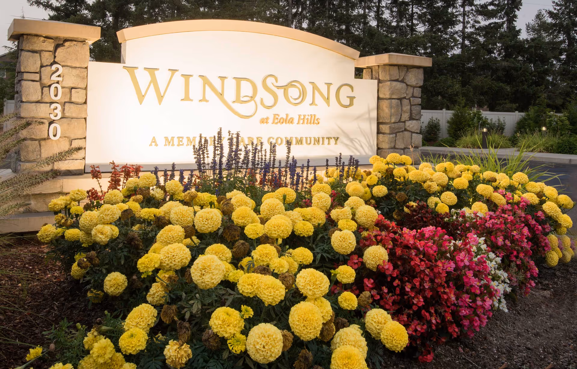 Stone and white sign for Windsong at Eola Hills, a memory care community, surrounded by vibrant yellow and red flowers with trees and a white fence in the background.