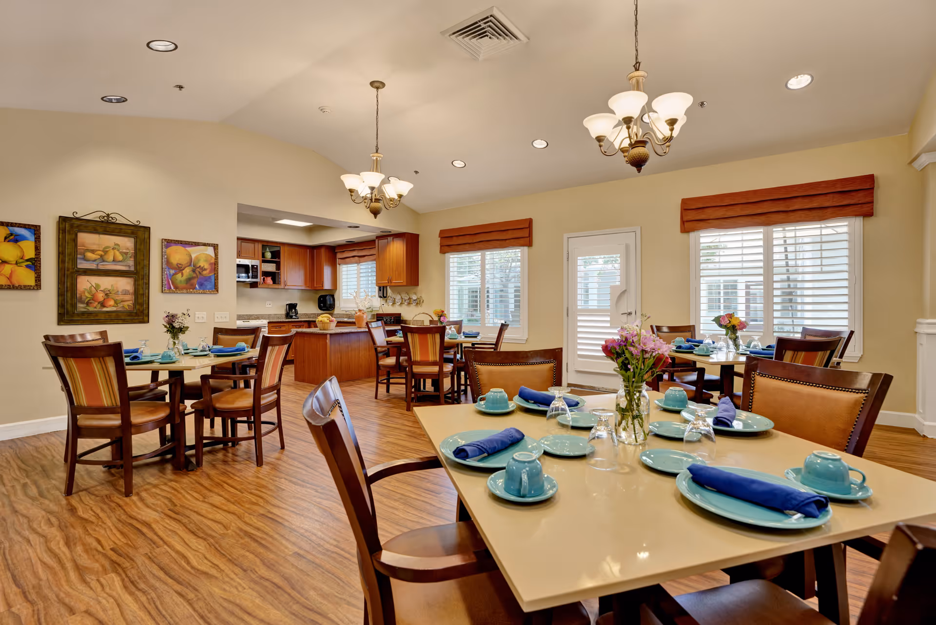 A bright dining room with several tables set with blue plates, cups, and napkins. The room features wooden chairs, hardwood flooring, and two chandeliers hanging from the ceiling. There are windows with blinds and a door leading outside. The background shows a kitchen area with wooden cabinets and appliances. The walls are decorated with framed fruit paintings.