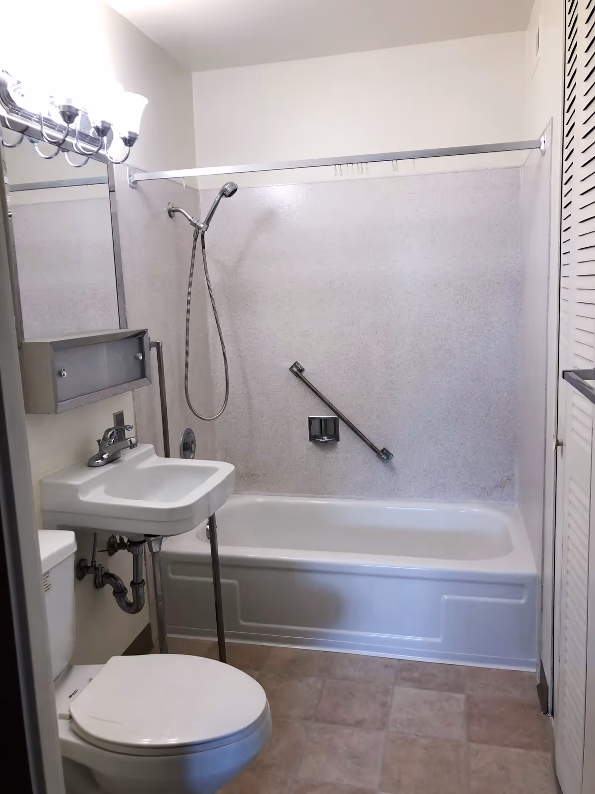 A small bathroom with a white toilet, a wall-mounted sink with exposed plumbing, a bathtub with a handheld showerhead, a grab bar on the wall, and a light fixture above a mirror.
