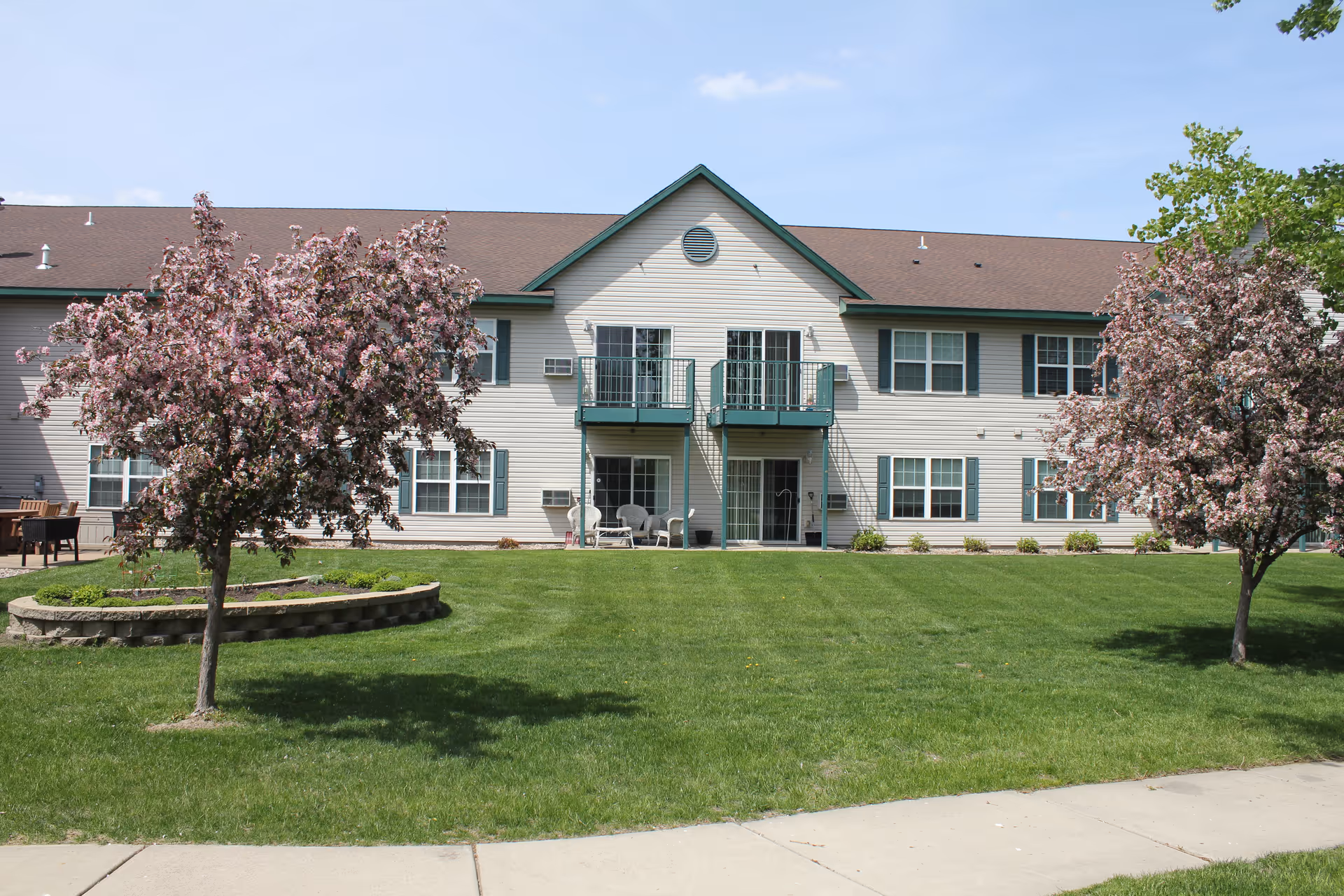 Exterior view of a two-story senior living facility building with beige siding and green trim. The building has balconies on the second floor and a well-maintained green lawn in front with two blooming pink flowering trees. A concrete sidewalk runs along the front of the lawn under a clear blue sky.