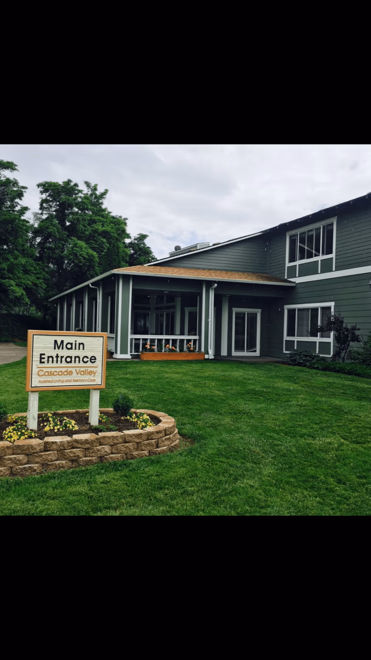 Exterior view of a green two-story building with white trim, identified as Cascade Valley Assisted Living and Memory Care. There is a well-maintained lawn and a flower bed with a sign that reads 'Main Entrance Cascade Valley Assisted Living and Memory Care'. The sky is overcast and there are trees in the background.