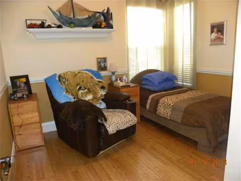 A bedroom with two beds, a chair, dresser, and window blinds in a senior care home.