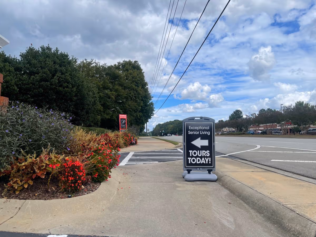 Sidewalk and roadside sign reading 'Exceptional Senior Living — Tours Today!' next to flower beds and a wide street under a cloudy sky.