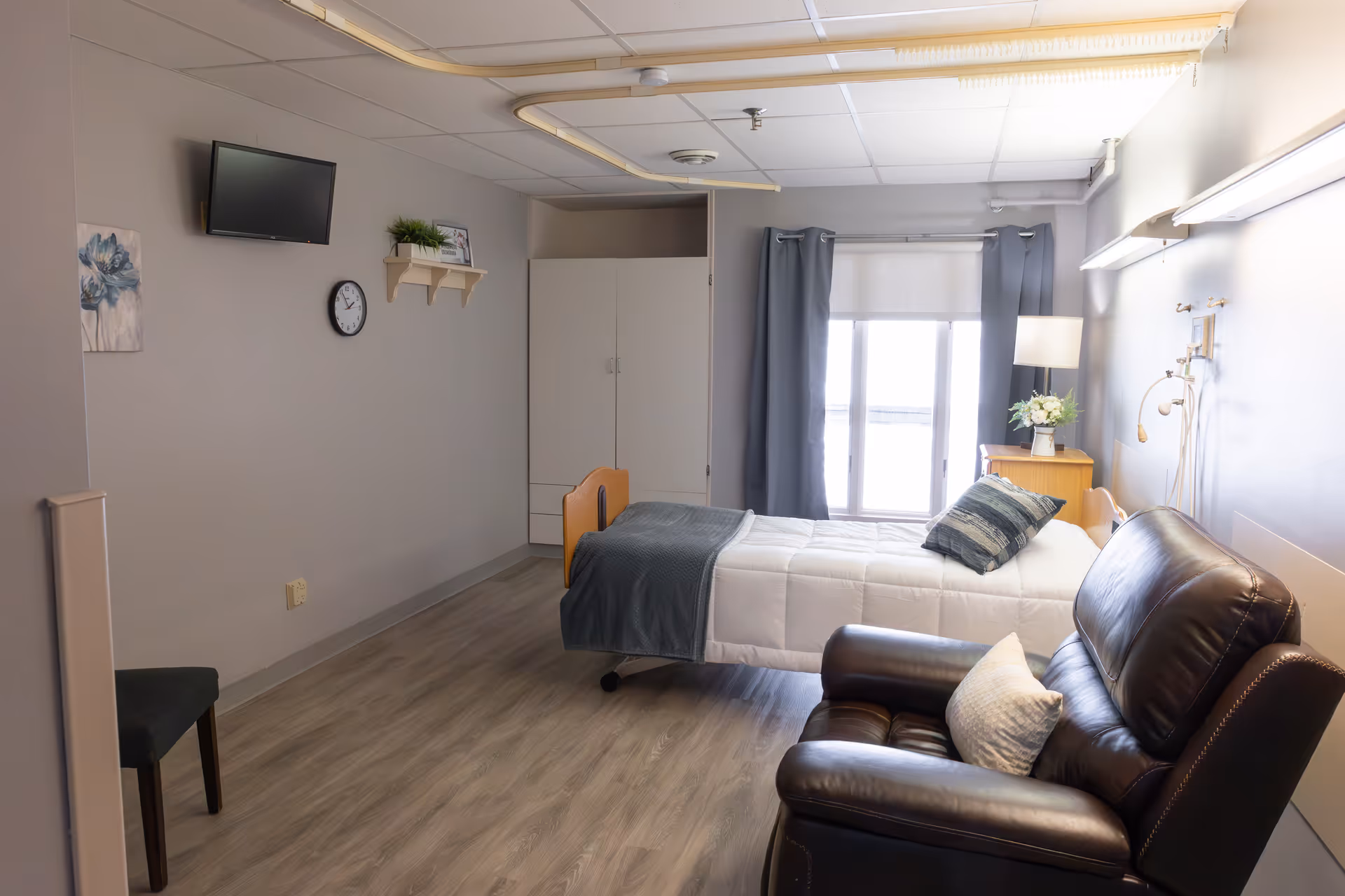 A clean and well-lit bedroom in a senior living facility featuring a single bed with white and gray bedding, a dark leather recliner chair with a pillow, a small wooden nightstand with a lamp and flowers, a wall-mounted TV, a clock, a small shelf with a plant and picture frame, gray curtains covering a window, and light wood flooring.