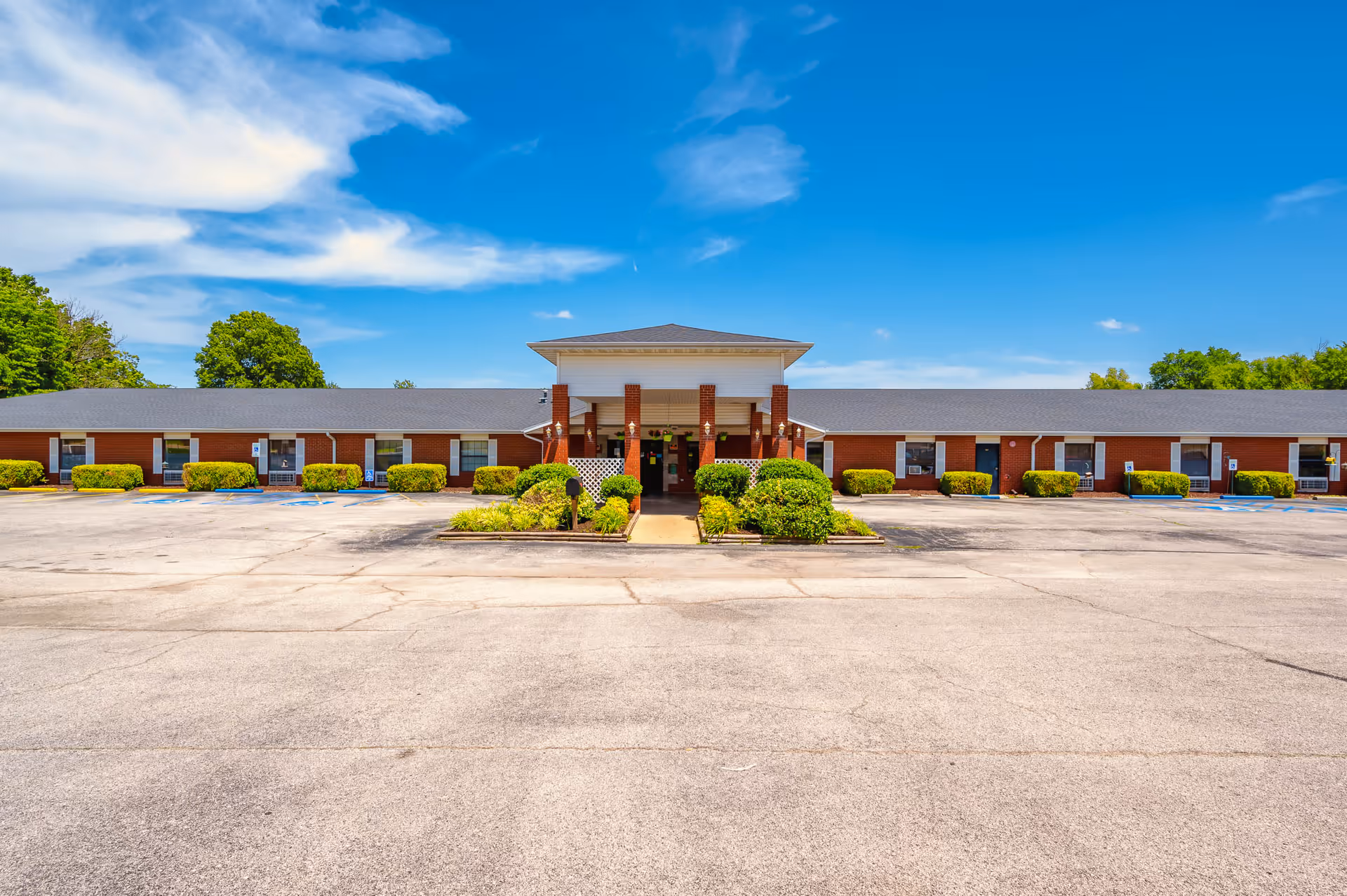 Front exterior of a single-story red-brick care facility with a covered entrance, landscaped shrubs, and a large empty parking lot under a blue sky.
