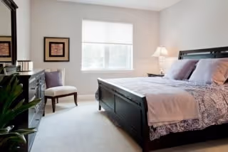 Well-lit bedroom with a dark wood bed, matching dresser, a chair, and a window with a shade.