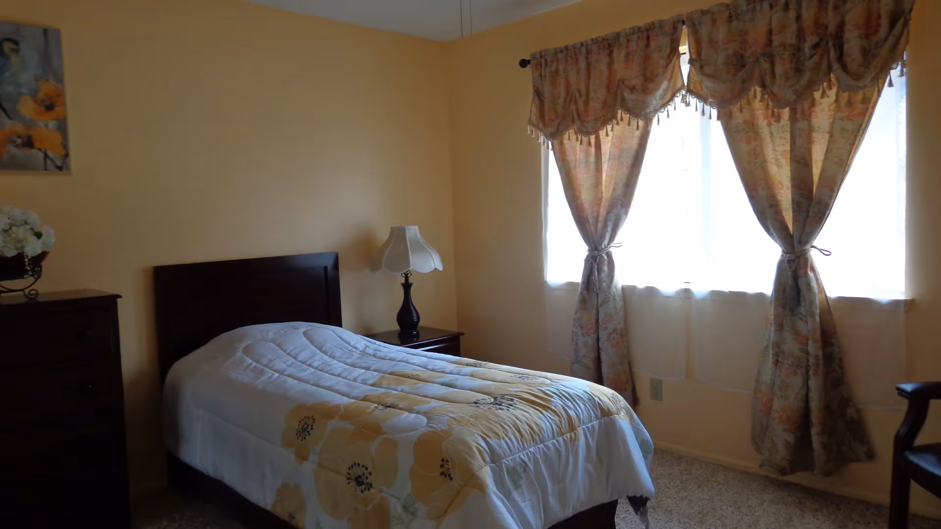 Small bedroom with a single bed, floral bedspread, nightstand with lamp, dresser and curtained window.