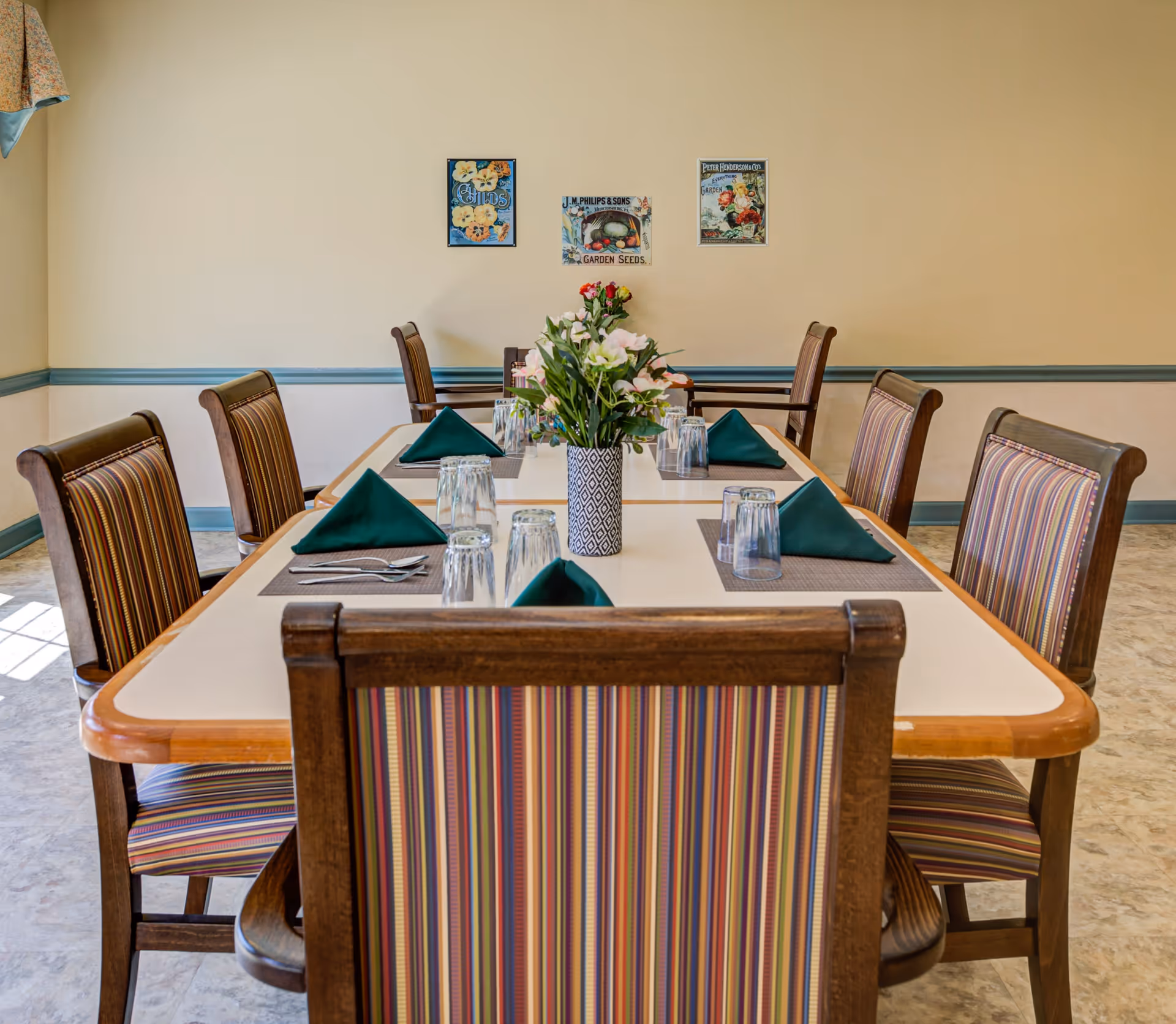 A dining room with a rectangular table set for six people. The table has green folded napkins, upside-down drinking glasses, and silverware on placemats. A vase with flowers is placed in the center of the table. The chairs have wooden frames with striped upholstery. The walls are beige with a blue chair rail, and three framed vintage-style posters hang on the wall.