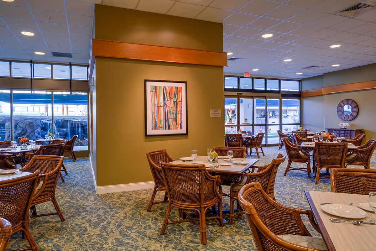 Dining room with wicker chairs and set tables, a green accent wall with framed artwork, and large windows.
