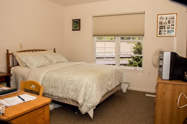 A bedroom with a neatly made bed featuring light-colored bedding. There is a wooden desk with a chair in the foreground, a window with a partially closed blind, and a wooden dresser with a television on top. The walls are light-colored and decorated with framed pictures.
