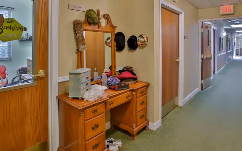 A hallway in a senior living facility with a wooden vanity table and mirror labeled 'SALON'. Various hats hang on the wall beside the mirror. The hallway has beige walls, green carpet, and several closed wooden doors. An exit sign is visible at the end of the hallway.