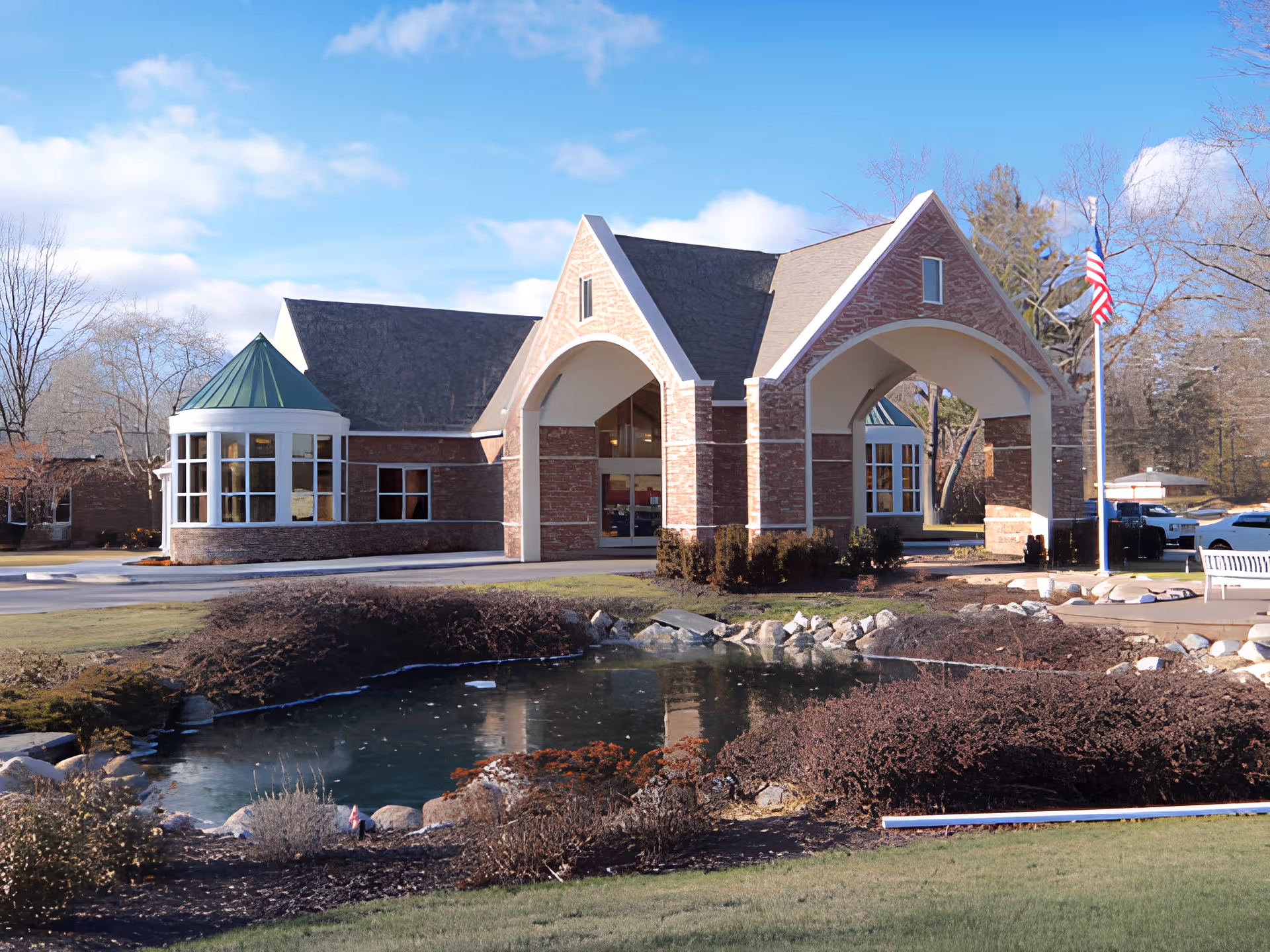 Exterior view of Manoogian Manor, a brick building with a large covered entrance supported by arches. The building has a green conical roof section and multiple windows. In front of the building is a small pond surrounded by rocks and landscaping, with an American flag on a flagpole to the right.