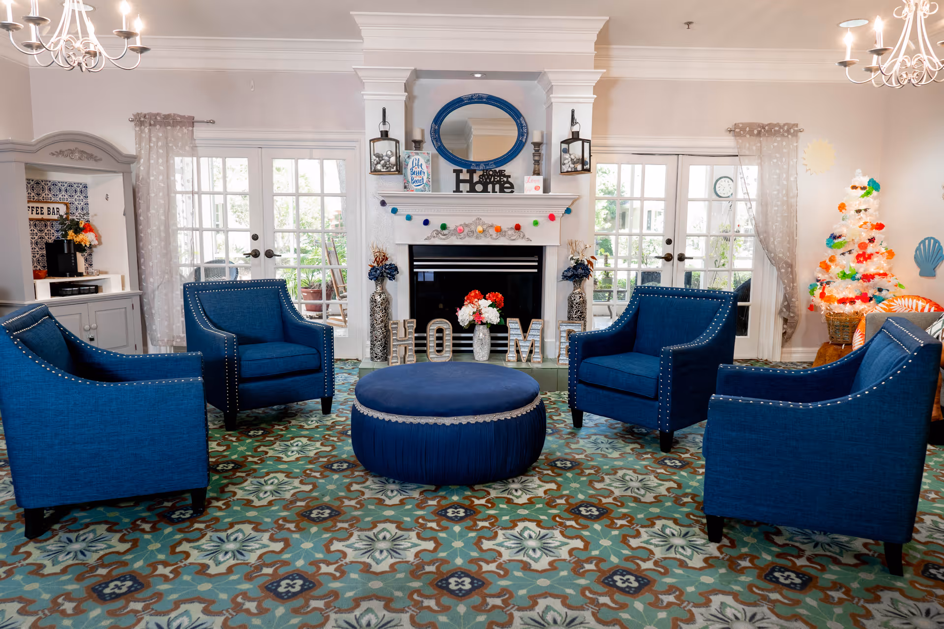 A cozy living room with four blue armchairs arranged around a round blue ottoman on a patterned carpet. The room features a white fireplace decorated with a blue oval mirror, lanterns, and decorative letters spelling 'HOME'. There are double glass doors on either side of the fireplace letting in natural light. A small decorated Christmas tree is visible on the right side, and a coffee bar area is on the left.