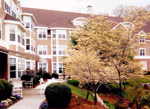 Exterior view of a multi-story senior living building with a landscaped courtyard, walkway, patio seating, and trees.