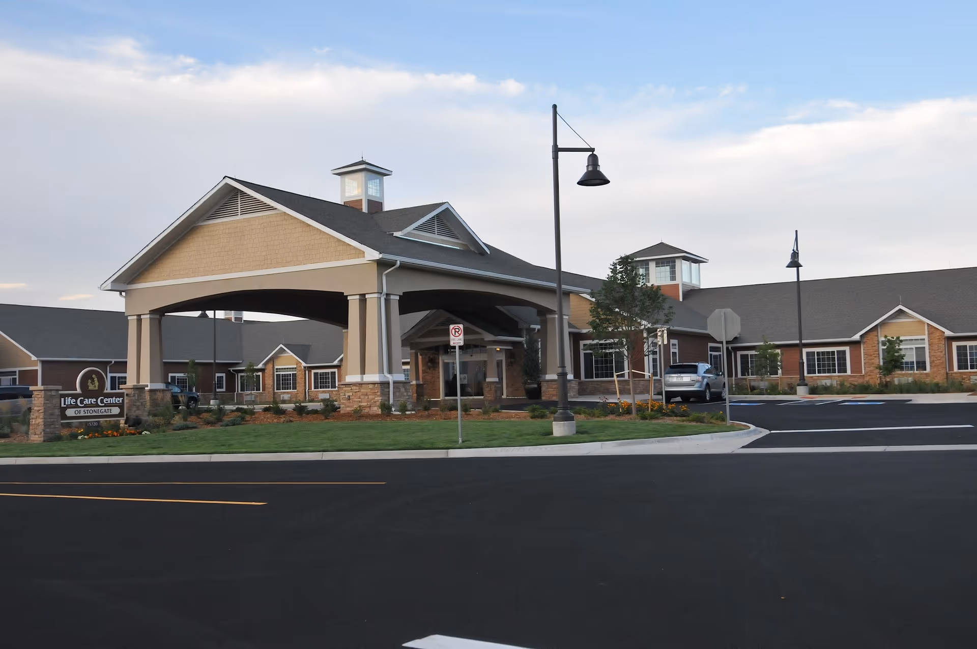 Exterior view of the Life Care Center of Stonegate building with a covered entrance, parking lot, street lamps, and landscaped greenery under a partly cloudy sky.